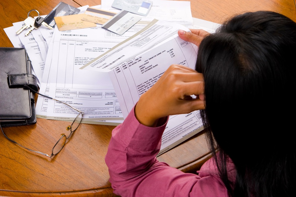 A woman looking at bills, with credit cards, glasses, and a notebook on the table